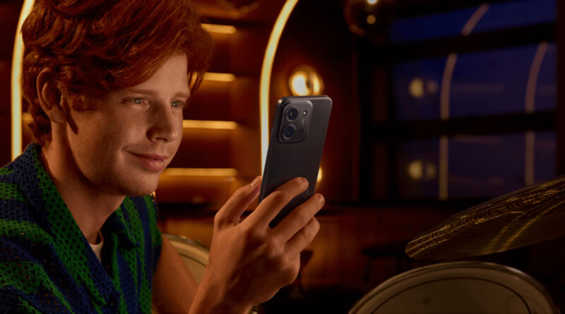 Joven con cabello rojizo sosteniendo un smartphone azul en un ambiente iluminado con tonos cálidos y detalles dorados.