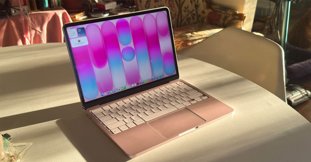 MacBook en color rosa claro con fondo de pantalla en tonos rosa y azul, sobre una mesa iluminada por la luz del sol.