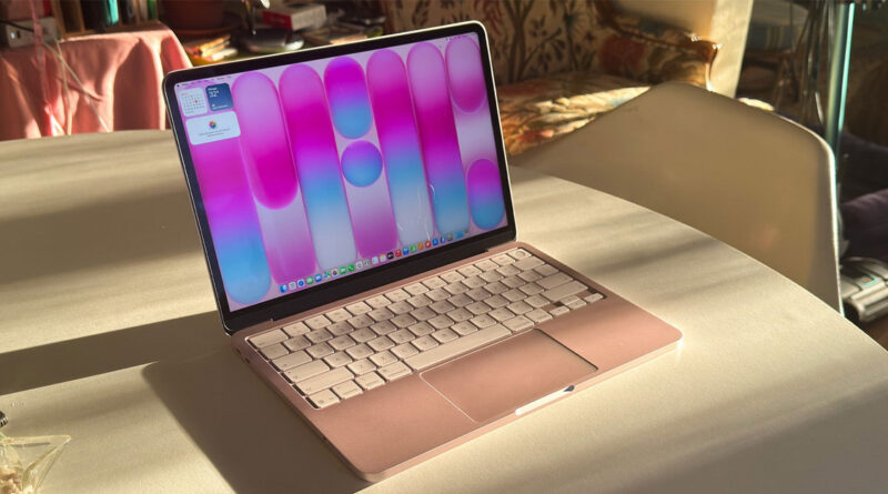 MacBook en color rosa claro con fondo de pantalla en tonos rosa y azul, sobre una mesa iluminada por la luz del sol.