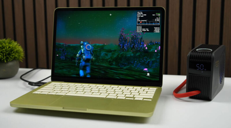 Laptop MacBook con pantalla de juego en un entorno alienígena, junto a un dispositivo de enfriamiento en una mesa blanca.