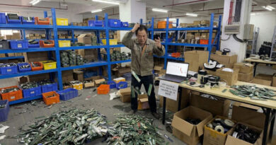 Trabajador angustiado en almacén, montones de módulos RAM tirados en el suelo, estanterías azules y laptop mostrando gráfico de caída de precios.