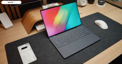 Laptop Dell XPS 14 sobre una mesa de madera, con pantalla colorida, teclado plano y un ratón blanco a su lado. Fondo minimalista.