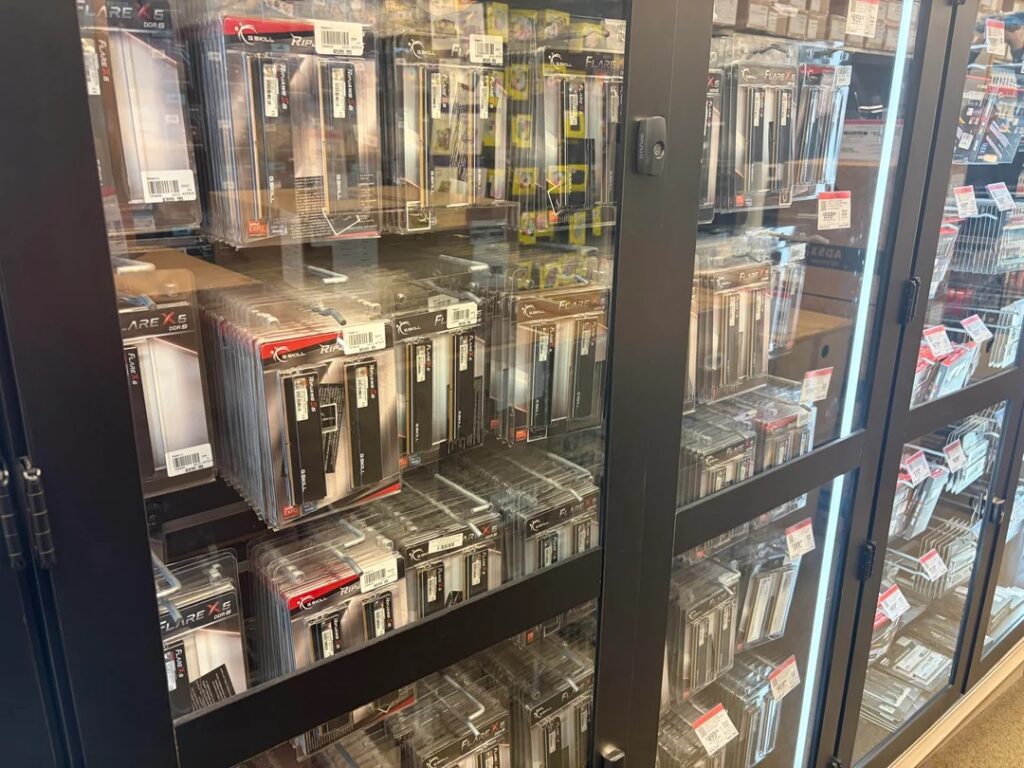 Estantes de vidrio repletos de paquetes de RAM y SSD, con etiquetas de precios y colores variados, en un entorno de tienda.