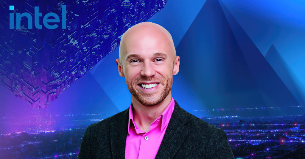 Hombre sonriente con cabello rapado, vestido con camisa rosa y chaqueta oscura, frente a un fondo tecnológico azul y púrpura.