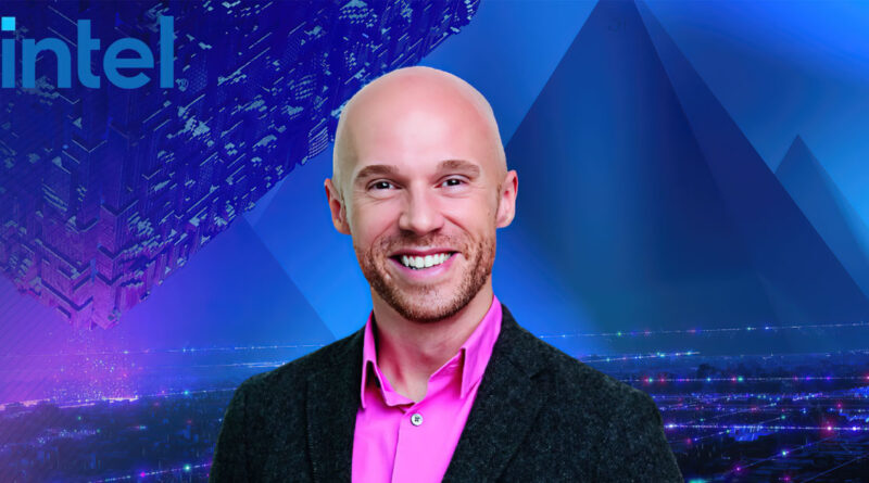 Hombre sonriente con cabello rapado, vestido con camisa rosa y chaqueta oscura, frente a un fondo tecnológico azul y púrpura.