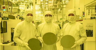 Tres trabajadores en trajes de protección sostienen obleas en una fábrica iluminada con luz amarilla, rodeados de maquinaria.