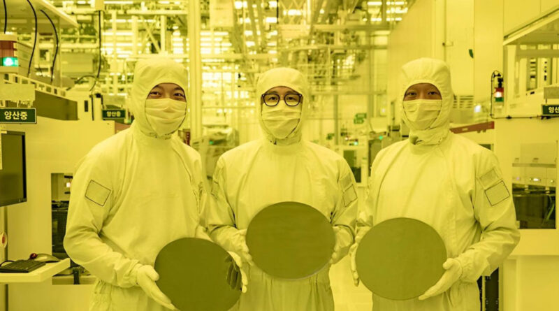 Tres trabajadores en trajes de protección sostienen obleas en una fábrica iluminada con luz amarilla, rodeados de maquinaria.