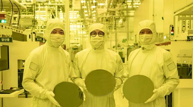 Tres trabajadores en trajes de protección sostienen obleas en una fábrica iluminada con luz amarilla, rodeados de maquinaria.