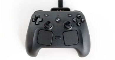 Controlador negro con joysticks, botones y superficie texturizada, conectado a un puerto USB en una base. Fondo blanco.