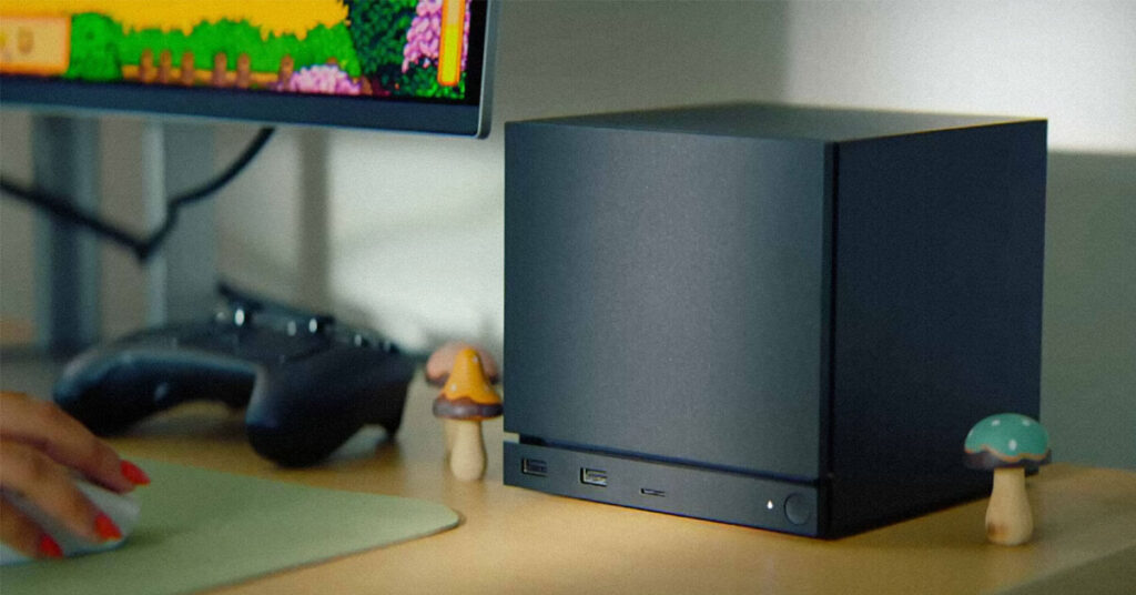 Computadora compacta negra sobre un escritorio, con un monitor que muestra un juego retro y decoraciones de hongos a los lados.