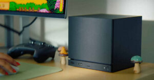 Computadora compacta negra sobre un escritorio, con un monitor que muestra un juego retro y decoraciones de hongos a los lados.