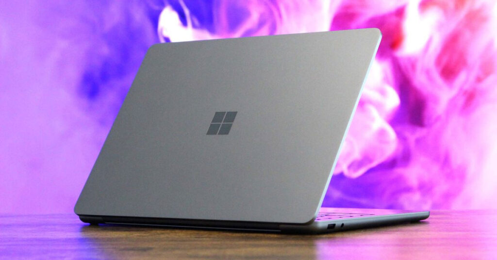 Laptop gris mate de Microsoft con logotipo en el centro, apoyada sobre una superficie de madera. Fondo con humo morado.