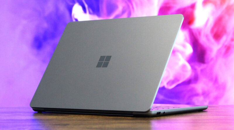 Laptop gris mate de Microsoft con logotipo en el centro, apoyada sobre una superficie de madera. Fondo con humo morado.