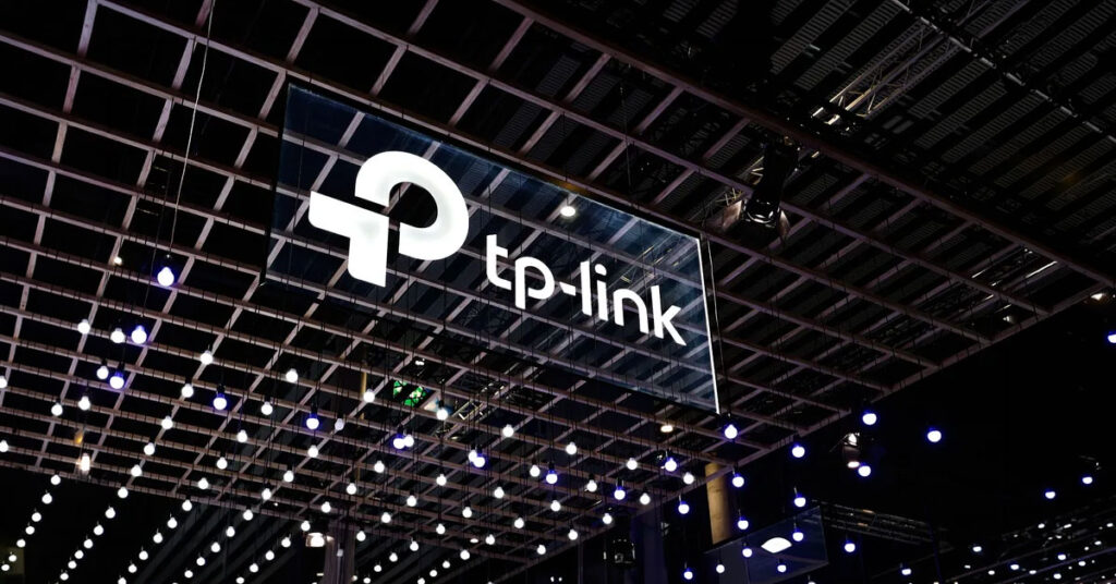 Logotipo de TP-Link suspendido en un espacio iluminado con luces colgantes, sobre un fondo oscuro de una feria tecnológica.