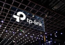 Logotipo de TP-Link suspendido en un espacio iluminado con luces colgantes, sobre un fondo oscuro de una feria tecnológica.