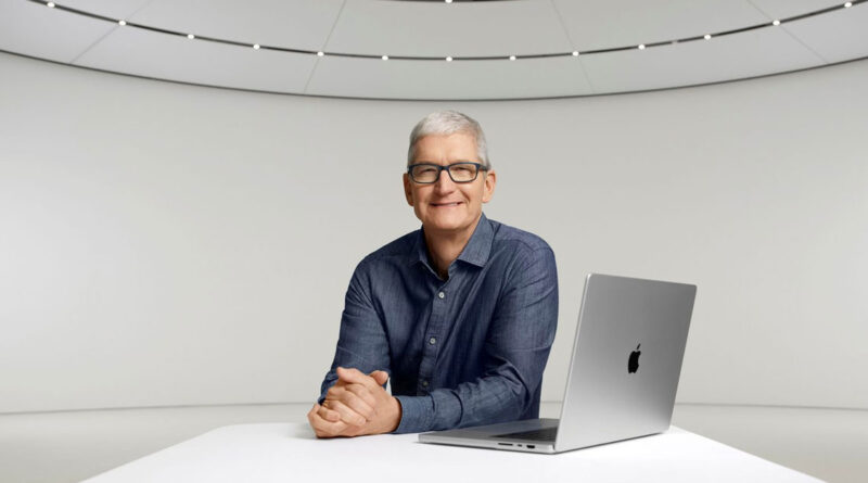 Hombre con cabello canoso sonriente, sentado en una mesa blanca con una laptop de Apple, fondo minimalista y de color claro.