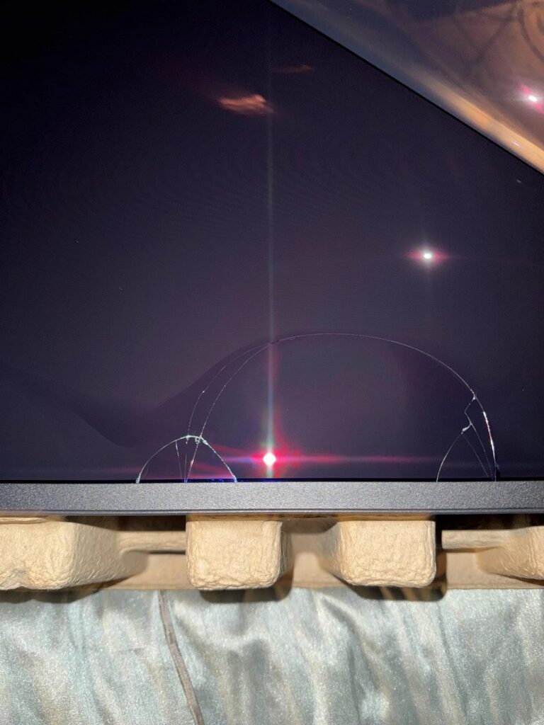 Pantalla de monitor con una fractura visible en la esquina inferior, sobre un fondo de embalaje de cartón y textura suave.