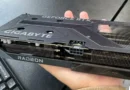 Tarjeta gráfica Gigabyte RTX 5060 Ti con disipador de Radeon, sostenida en una mano, en un fondo de mesa oscura.