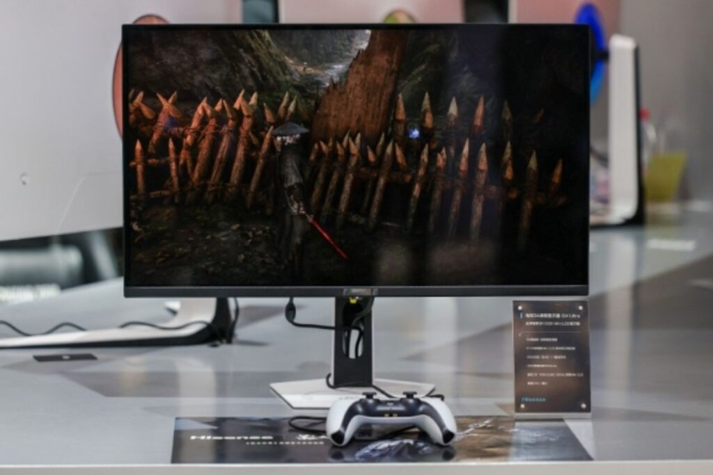 Monitor de 5K con una imagen de juego brillante, soporte negro, y un controlador de PlayStation en primer plano. Colorido y moderno.
