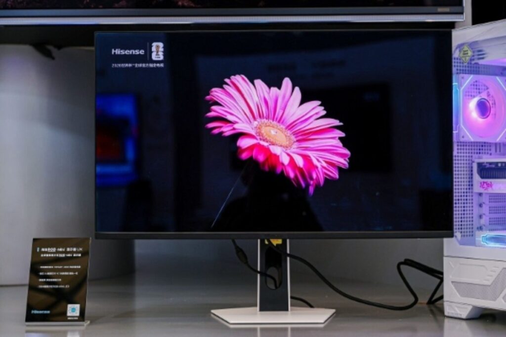 Monitor Hisense con pantalla grande mostrando una flor rosa vibrante, soporte blanco y PC de juegos iluminada a su lado.