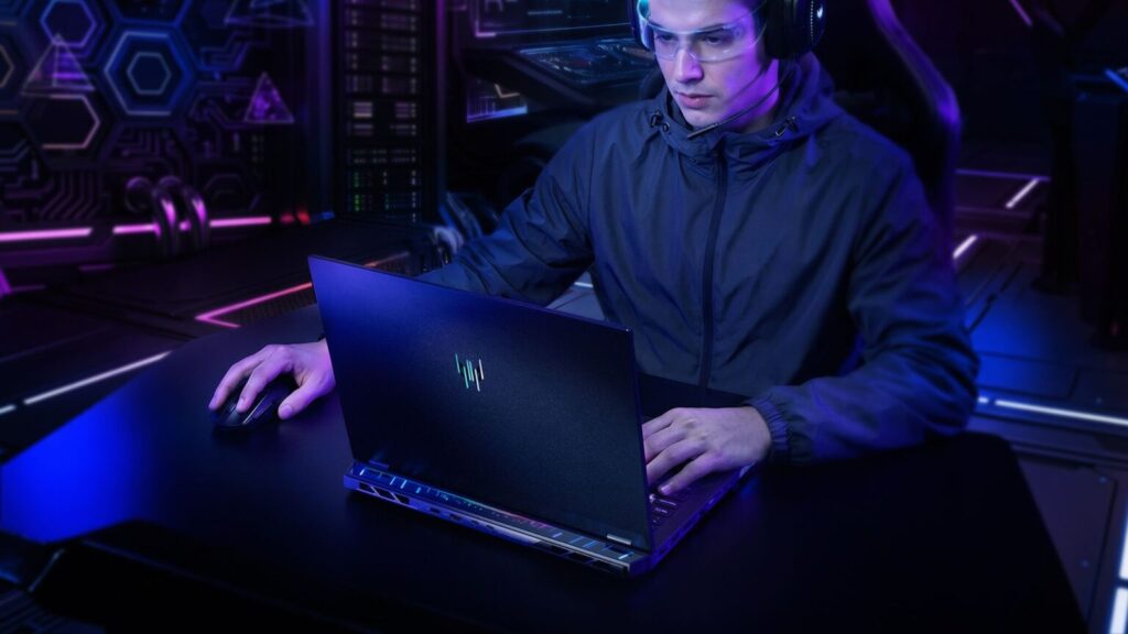 Joven con auriculares y gafas usando una laptop de gaming oscura con luces, sentado frente a un fondo tecnológico iluminado.