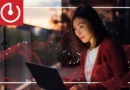 Mujer usando una laptop al atardecer, con ondas rojas de energía alrededor y un logotipo de encendido en la esquina superior izquierda.