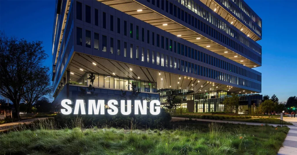 Edificio moderno de Samsung iluminado en la noche, con un gran logotipo blanco en la entrada y vegetación en primer plano.