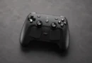 Controlador negro de videojuegos con botones y joysticks, sobre una superficie gris oscura, resaltando su diseño moderno y minimalista.