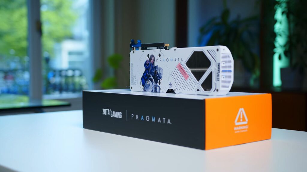 Tarjeta gráfica Zotac RTX 5070 AMP PRAGMATA, con diseño en colores blanco y azul, presentada sobre una caja negra y naranja.