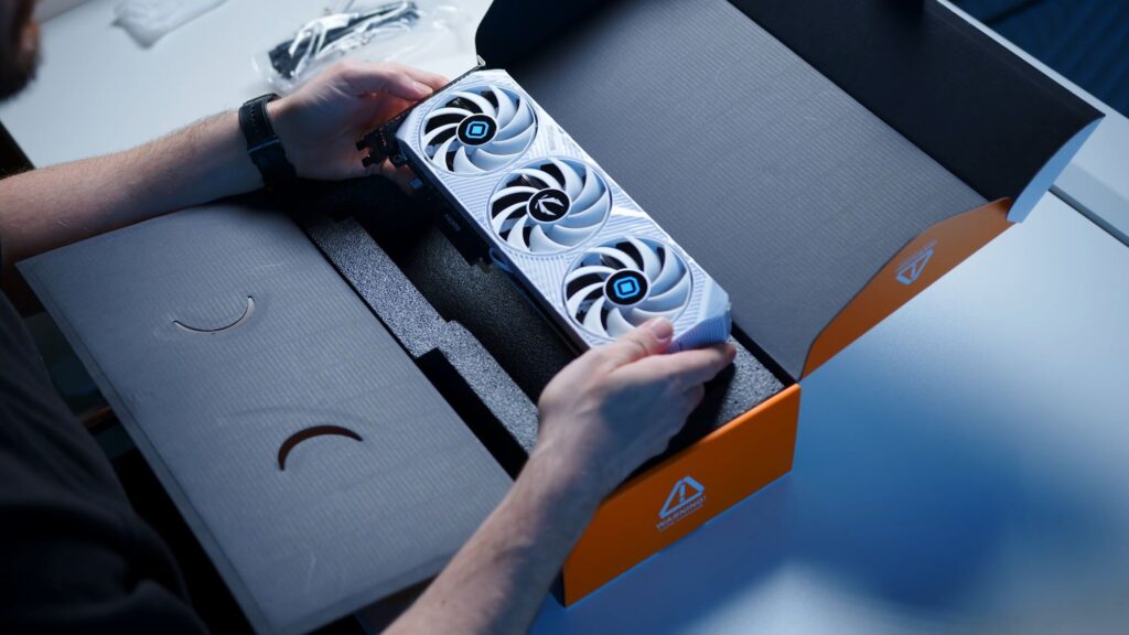 Tarjeta gráfica RTX 5070 AMP PRAGMATA en una caja abierta, sostenida por manos, con un diseño blanco y ventiladores destacados en negro y azul.