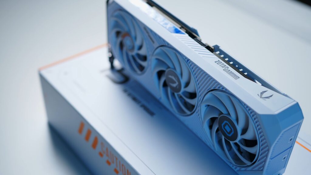 Tarjeta gráfica RTX 5070 AMP PRAGMATA en tonos blanco y azul, con tres ventiladores y un diseño moderno, sobre una caja blanca.