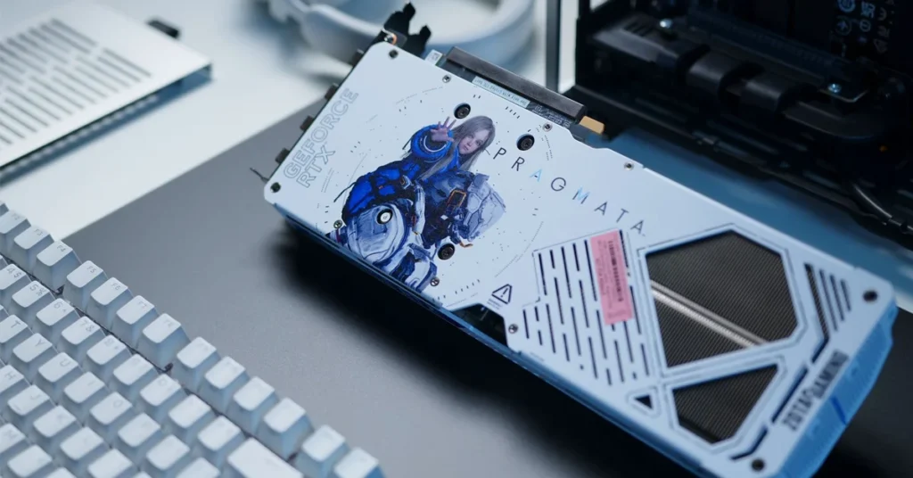 Tarjeta gráfica Zotac RTX 5070 AMP PRAGMATA con diseño artístico de personajes, colores blanco y azul, sobre una superficie oscura con un teclado.