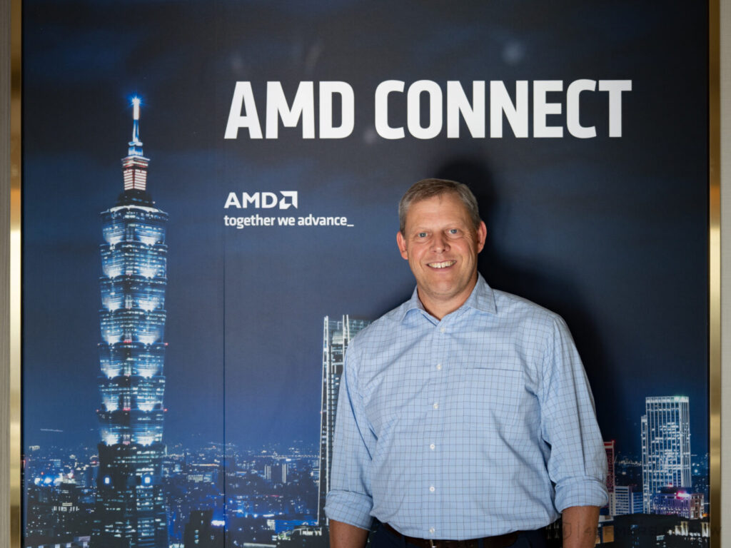 Hombre sonriente frente a un fondo de AMD Connect, con la ciudad iluminada de Taipéi y la Torre Taipei 101 visible.