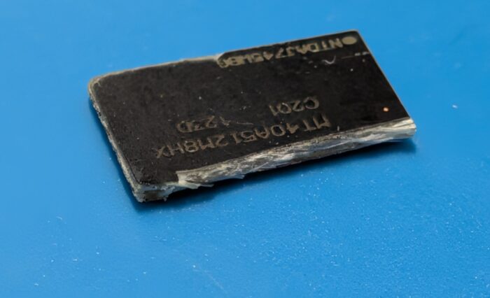 Chip de circuito negro con texto grabado en la superficie, sobre un fondo azul claro. Muestra signos de desgaste y daño.