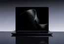 Portátil negro con pantalla oscura y suave curva en el fondo, sobre una superficie reflejante, en un entorno minimalista.