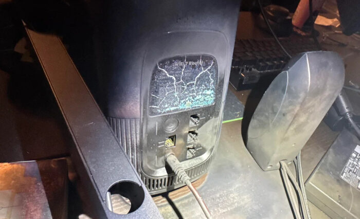 Router dañado con una pantalla agrietada, cables conectados y teclado al fondo; ambiente oscuro y desordenado.